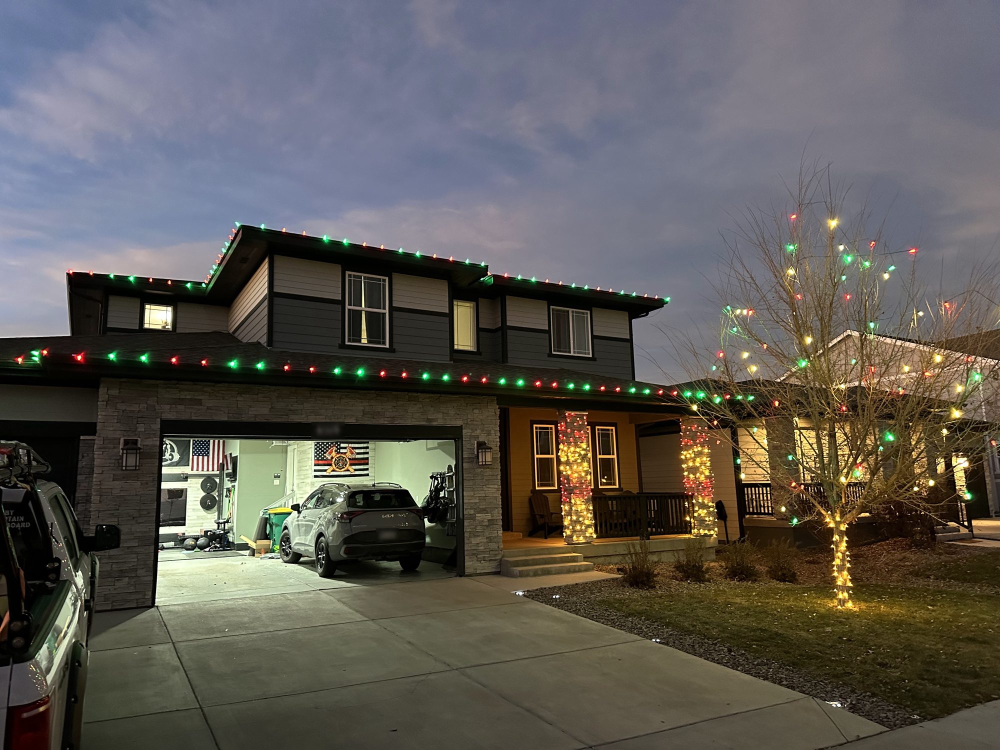 Two-story home at dusk with red and green roofline lights, warm white wrapped porch columns, and a multicolor-lit front yard tree